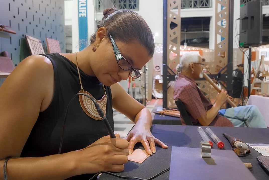 In the foreground, a woman with dark hair, glasses, and necklace works with an electric pen-like tool in one hand on a small square of tan bark. Behind her, a man with white hair sits back, playing a wooden flute.
