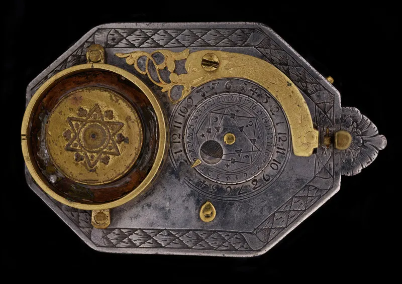 A pocket sundial, seen from below. The sundial is covered with both functional and decorative etchings as well as gold accents. This side includes a lunar volvelle and a golden piece in the shape of a dolphin.