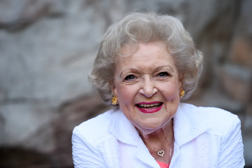 White, a white woman with curly white hair, smiles at the camera in a bright white suit with a heart shaped necklace