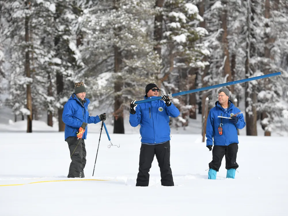 Three men in blue coats standing in the snow with equipment
