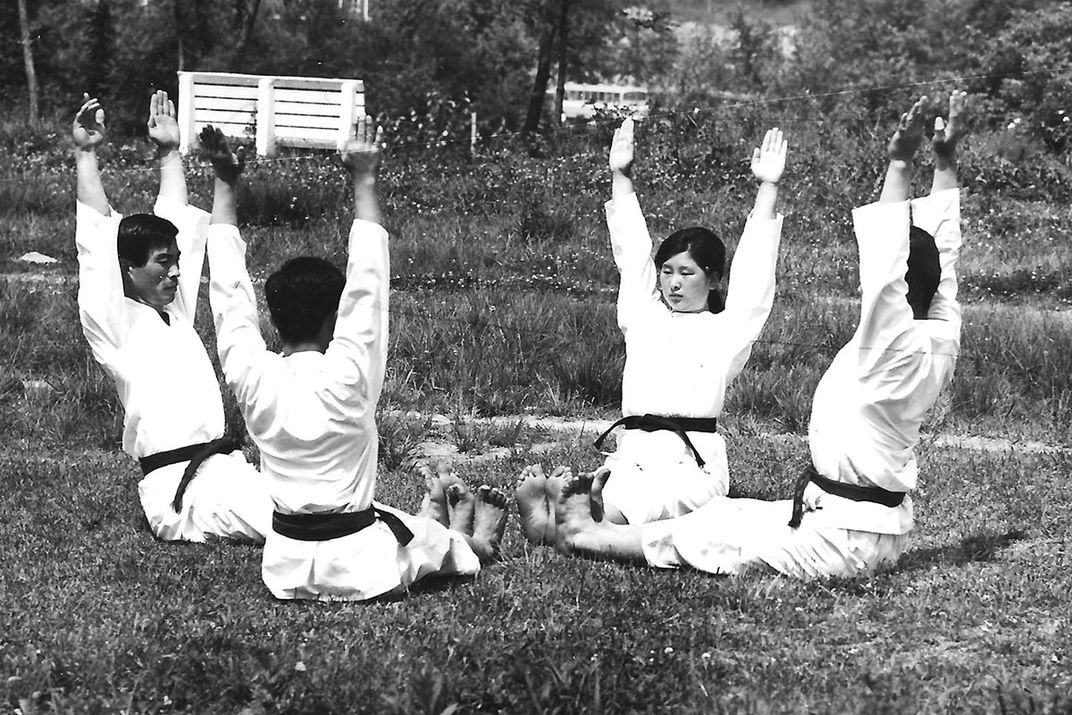 Four individuals sit in grass and raise their hands during a yoga demonstration.