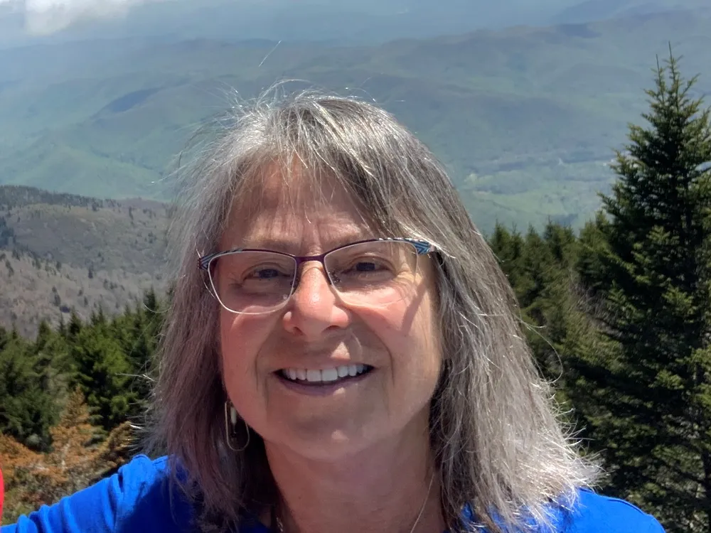 Head and shoulders photo of Denise Breitburg, a woman with glasses and shoulder-length gray hair, with forested mountains in the background.