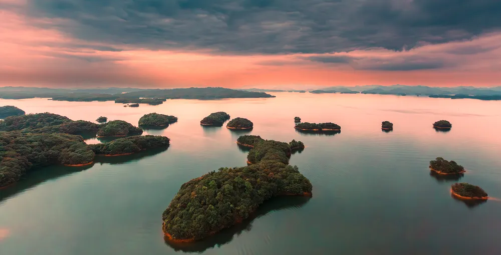 "When the lens focuses on Xihai(west sea), an exquisite landscape painting unfolds gently before the eyes. Lushan Xihai is a treasure bestowed by nature—a flowing poem between heaven and earth, a silent painting rendered by the universe.

The lake’s surface, clear as a mirror, shimmers under the caress of sunlight, like thousands of scattered diamonds glistening on the water. Over 8,000 islands are dotted across the vast expanse like emeralds inlaid upon the azure canvas or jade pieces scattered under starlight. The Heart-shaped Island resembles a romantic verse written by nature itself; its naturally formed silhouette, adorned with lush trees and blooming flowers, whispers tales of love and tenderness. The ""Lone Tree in the Sea"" stands like a solitary sage between sky and water, weathering wind and rain, watching clouds gather and drift, silently witnessing the passage of time.

Ascending the Xihai Star Glass Tower feels like stepping into the clouds. Beneath transparent glass floors and amidst drifting mist, the panoramic view of countless islands unfolds. And as the sun sets, a grand symphony of light and water begins. The sun, like a gentle painter, brushes soft hues across the lake, while distant mountains embrace the water in a tranquil ink-wash scene. Then, the horizon ignites with brilliant twilight, bursting like magnificent fireworks, setting the entire lake ablaze with ripples that shimmer like a fallen galaxy.

At Xihai, every drop of water carries the rhythm of poetry, every island holds the blank space of a painting, and every glow of sunset is nature’s heartfelt confession—leaving one enchanted, reluctant to depart."