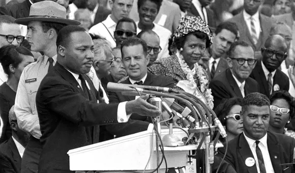 Martin Luther King, Jr. stands at a podium with several microphones. Behind him to his left is a crowd of seated and standing people, including Dorothy Height, turned towards him.