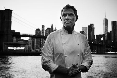 A black and white photograph of Bobby Flay in his signature white chef’s coat, with his initials, BF, embroidered on the front.