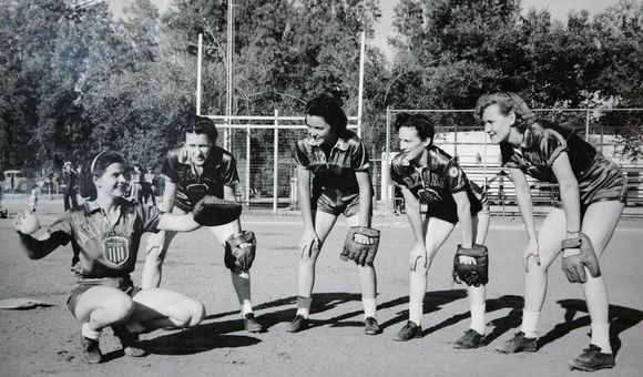 A group photo of women in softball uniforms wearing baseball gloves pose for a photo