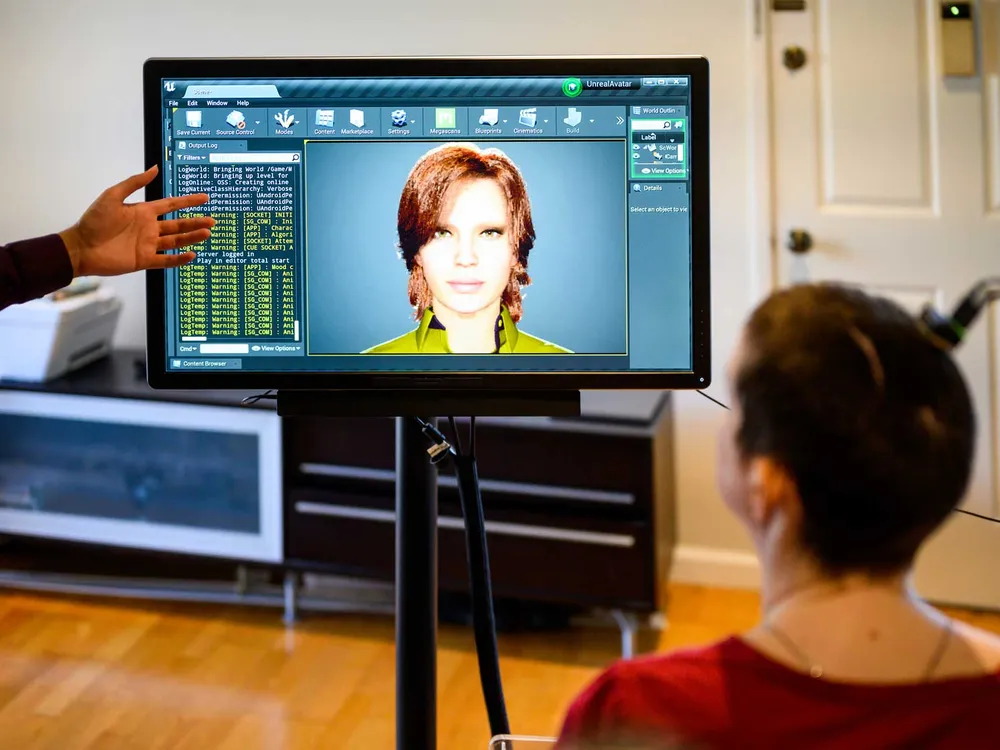 A woman sits facing a computer screen with a digital avatar on it