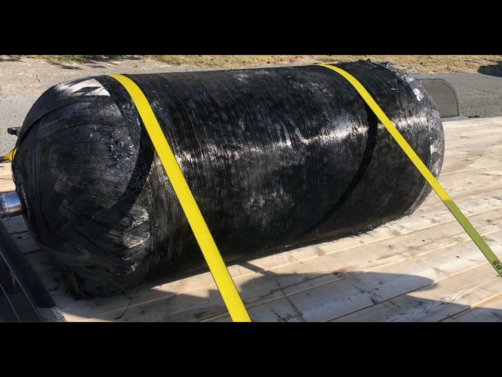 A photo of a large black cylinder strapped to a wood platform with yellow cords