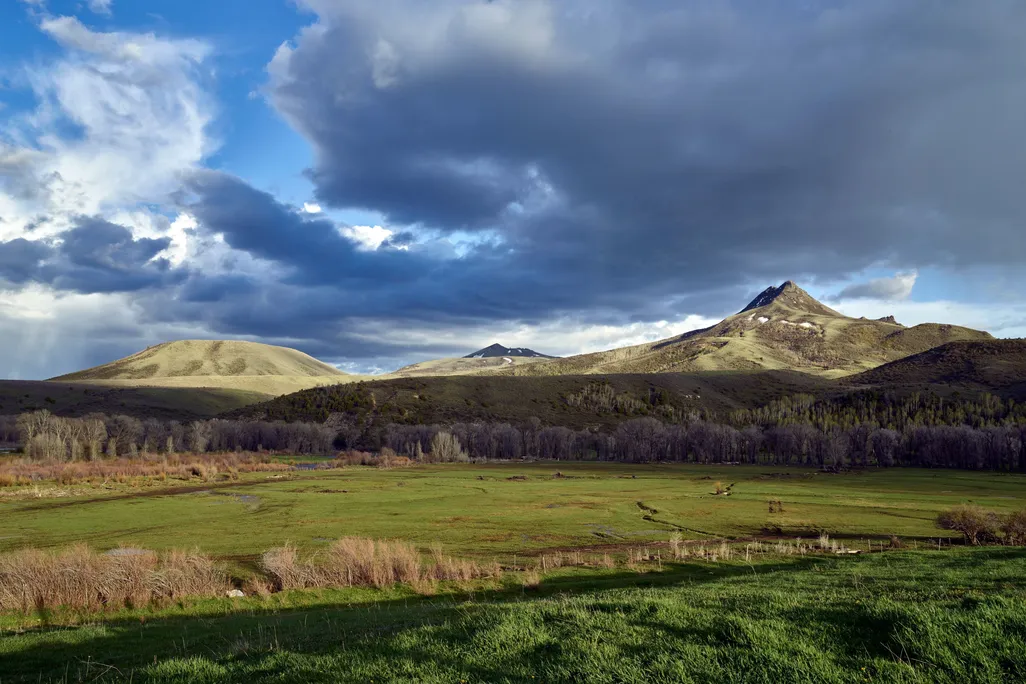 A view of a large field with a tall peak in right distance, framed by dramatic evening or morning light and large clouds crossing a blue sky