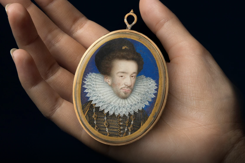 A white hand holds a small portrait in its palm; the oval frame is gilded and holds a portrait of a white man framed by blue, a starched ruffle around his neck and elaborate jewels