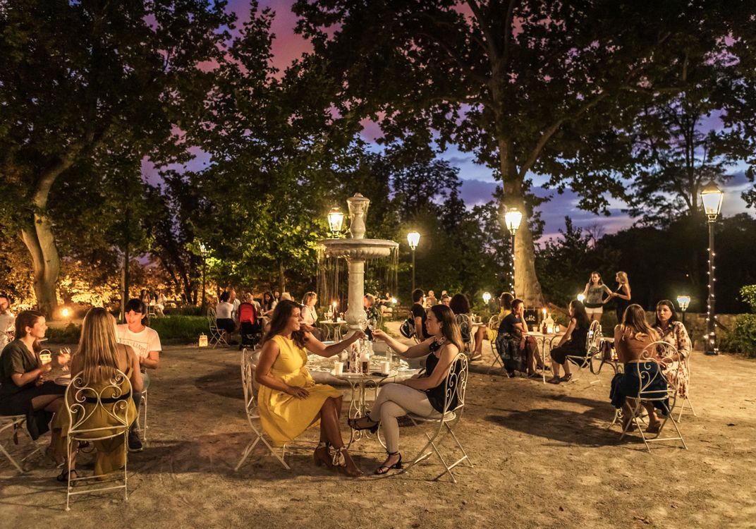 People seated at small outdoor tables around a fountain in a Zagreb park at dusk, with trees, lanterns, and soft lighting creating an inviting atmosphere.
