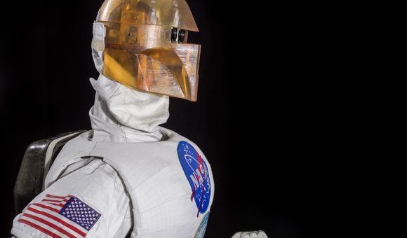 Photographed against a black background is a robot in profile wearing a silver uniform and golden-colored helmet.