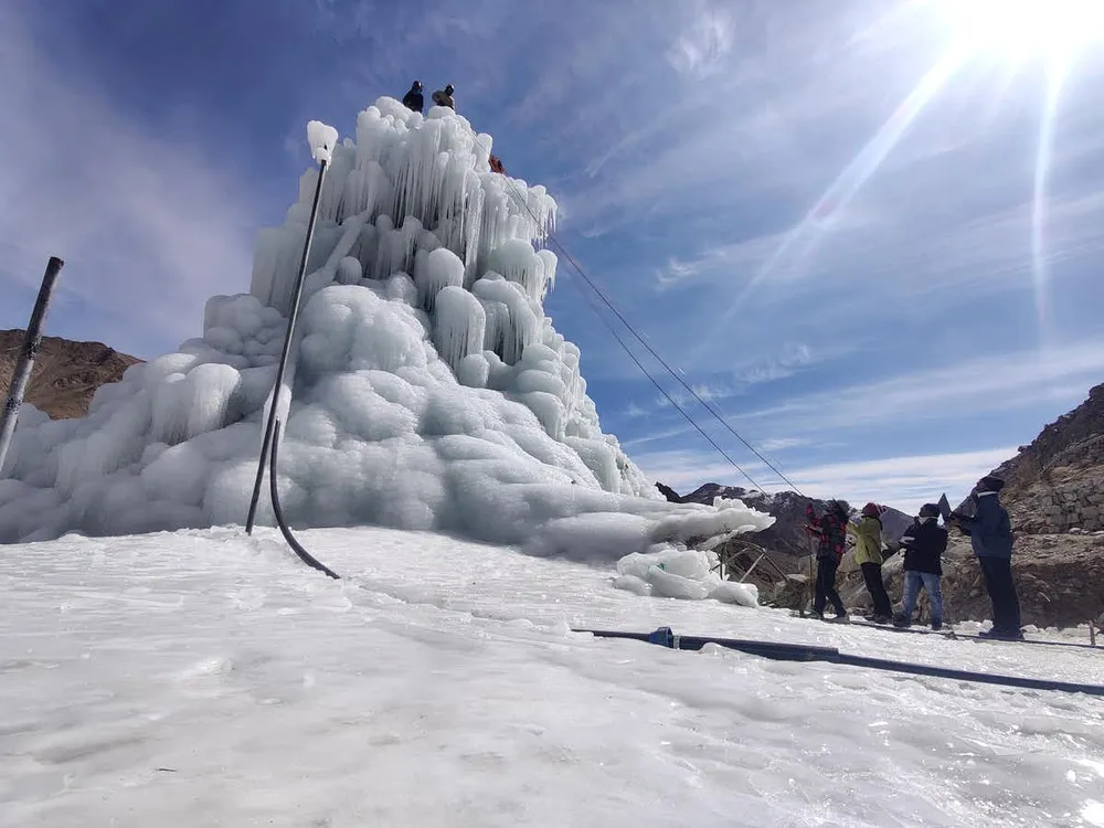 On a bright day in the Ladakh Valley, a team of people work on an ice stupa. It is a tall, cone-shaped pile of ice that sits upon snowy ground.