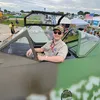 A white man in his 30s sits in the cockpit of a World War II-era military airplane. He is smiling and wearing sunglasses and a ballcap.