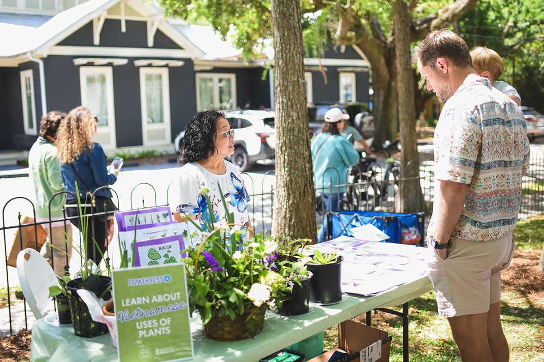 A woman with dark wavy hair speaks with a man across a table outdoors. Her table is set with potted plants and a sign that reads: Learn about Vietnamese uses of plants.