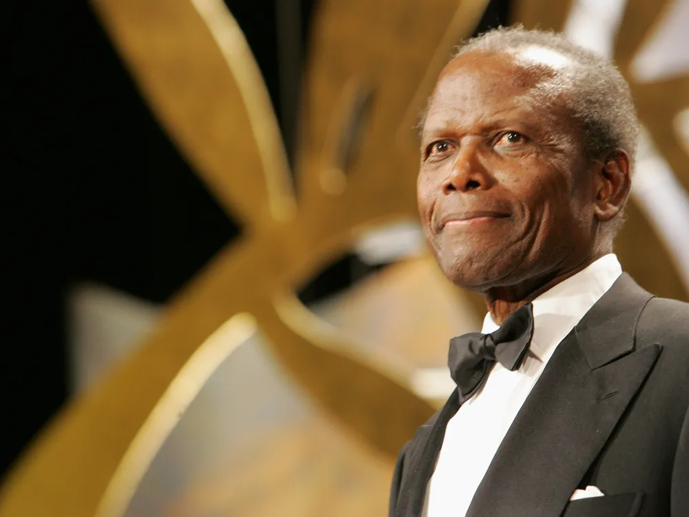 An elderly Poitier wears a black suit, white shirt and black bowtie and stands in front of a gold and silver background