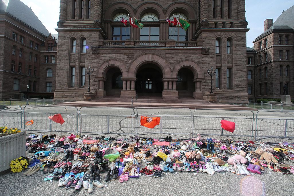 A memorial of children's shoes, all different colors, and flags are clustered on the concrete ground next to a fence, looking onto a big official building