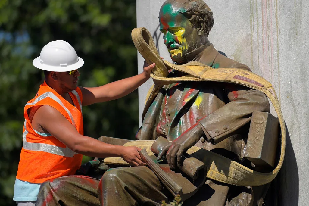 A man in a bright orange construction vest and white hat wraps a thick rope around a paint-splattered statue, of a bearded man wearing a suit and sitting in a chair