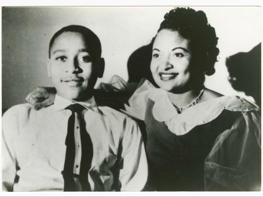 Emmett Till, a young boy in a suit and tie, smiles next to his mother, who wears a dress and has her hand on his shoulder