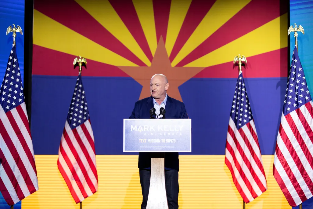 Mark Kelly stands on stage at a podium in front of the Arizona flag