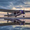 A vintage silver Ford Tri-Motor aircraft parked on a wet tarmac under a dramatic, cloudy sky at dusk. The plane features corrugated metal skin, three engines, and the "Ford" script logo in blue on the fuselage, all reflected in the water on the ground.