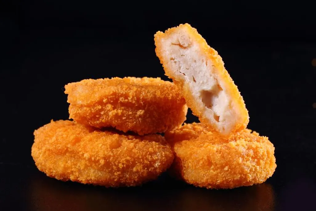 A close-up shot of three chicken nuggets stacked up in front of a black background. On top is one chicken nugget, cut in half, with the insides facing the camera. 