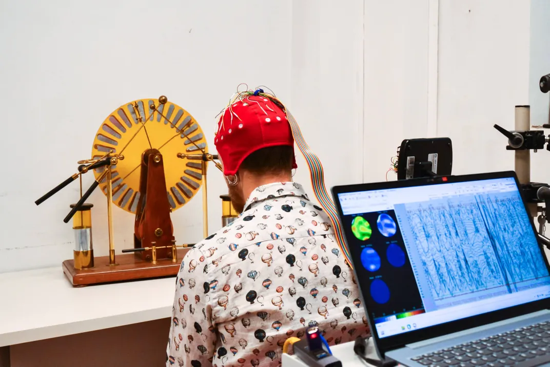 a person sits with his back to the camera facing a scientific instrument like an elevated wheel, a cap with electrodes is on his head and a computer screen shows brain activity
