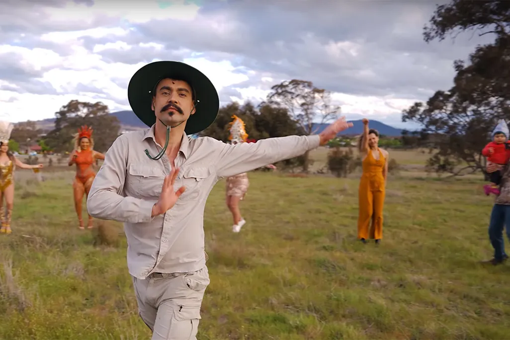 Weliton Menário Costa dances in the foreground of a grassy plain, while background dancers dressed in orange dance behind him.