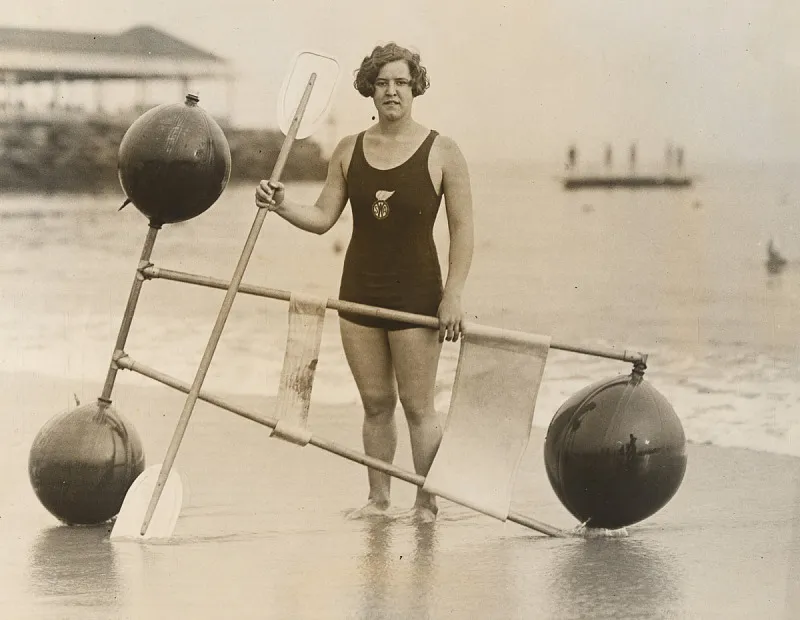 A black and white gelatin silver print of Gertrude Ederle standing on the shore with a 1920s flotation device.