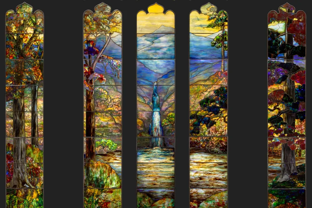 Five thin panels of stained glass, two on either side and three grouped closely together, depict a colorful landscape with blue mountain in the background and a waterfall flowing into a yellow green pond