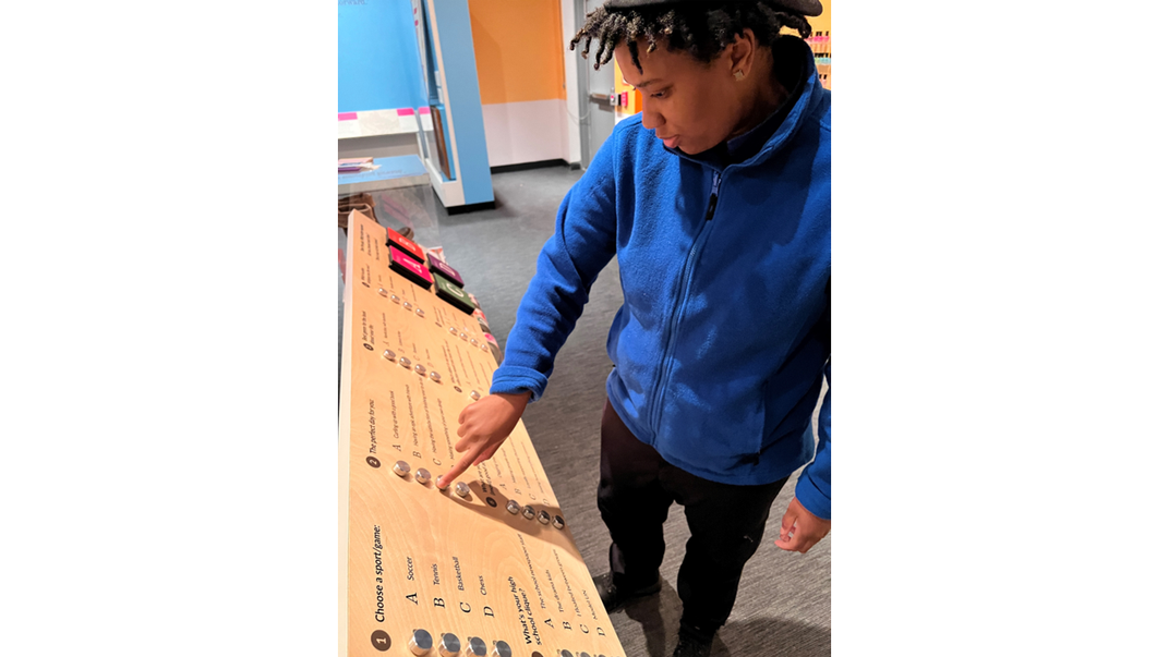 An African American young person is moving buttons on an interactive quiz board. The person is wearing a backward baseball cap, a blue fleece pull-over, and black pants.