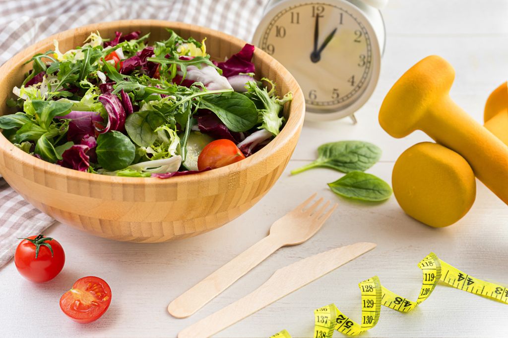 Wooden bowl of salad on a table with a clock, some utensils, a curled tape measure and barbells