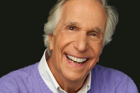 A headshot of Henry Winkler, shows a close-up the actor looking directly into the camera, his eyes twinkling above a broad, white smile.