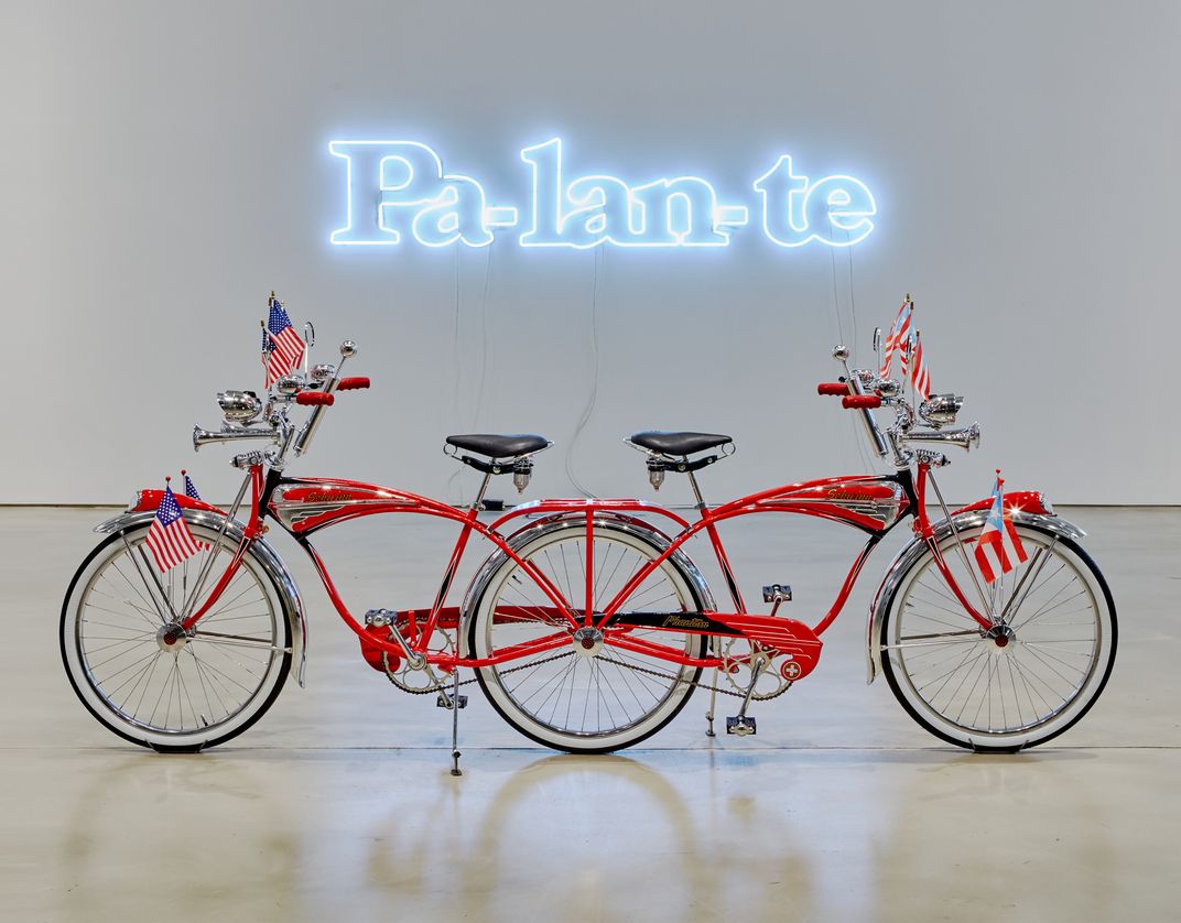 A sculpture of a red bicycle.  It has two front ends, joined together at the back.  Above, a neon light sculpture says "Pa-lan-te."