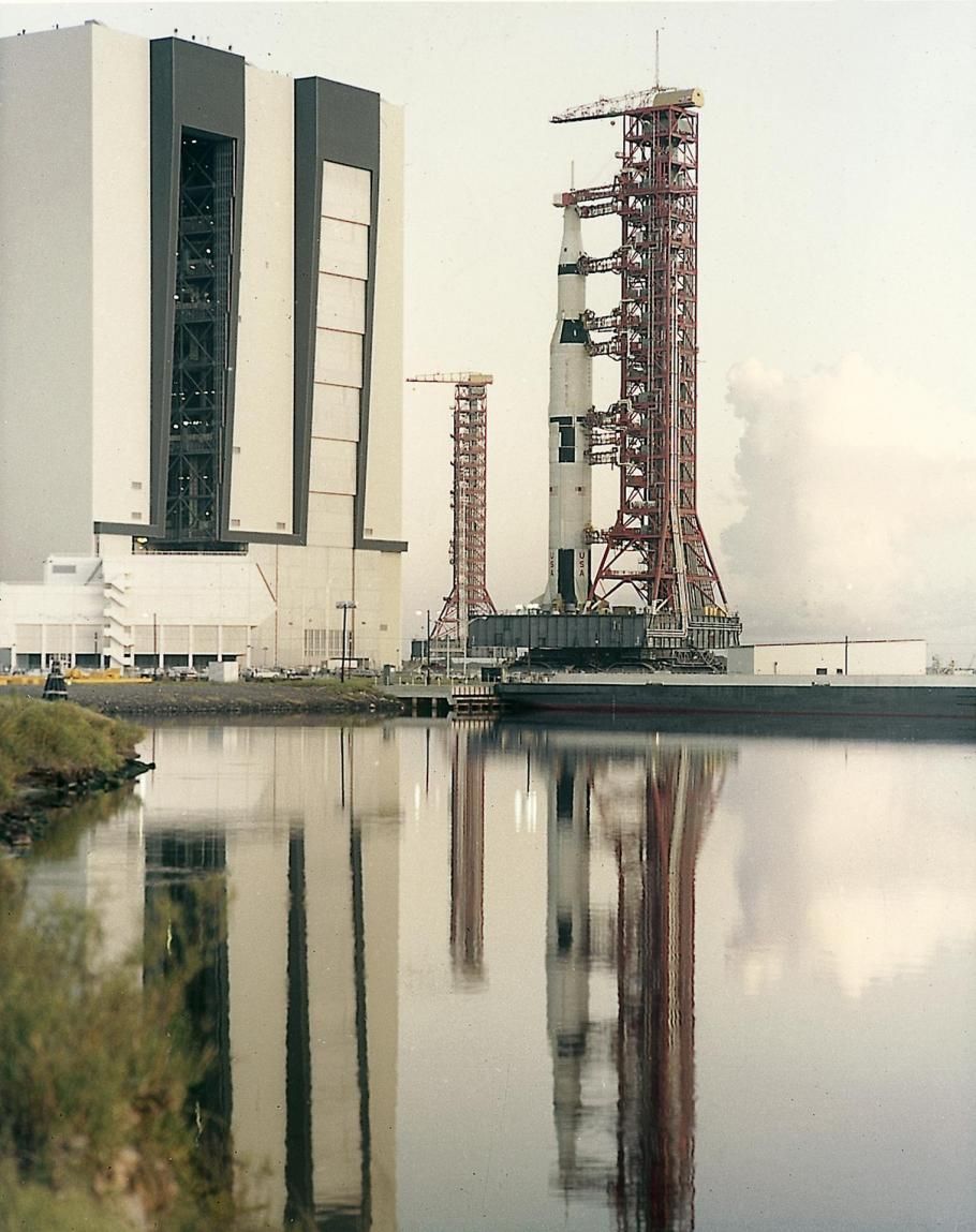 Saturn Five rocket on a mobile launcher outside the VAB. The rocket and mobile launcher are reflected in water beneath it.