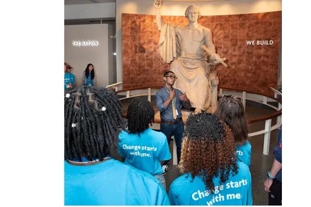 A group of students in bright blue shirts gather around a statue of George Washington with a museum facilitator
