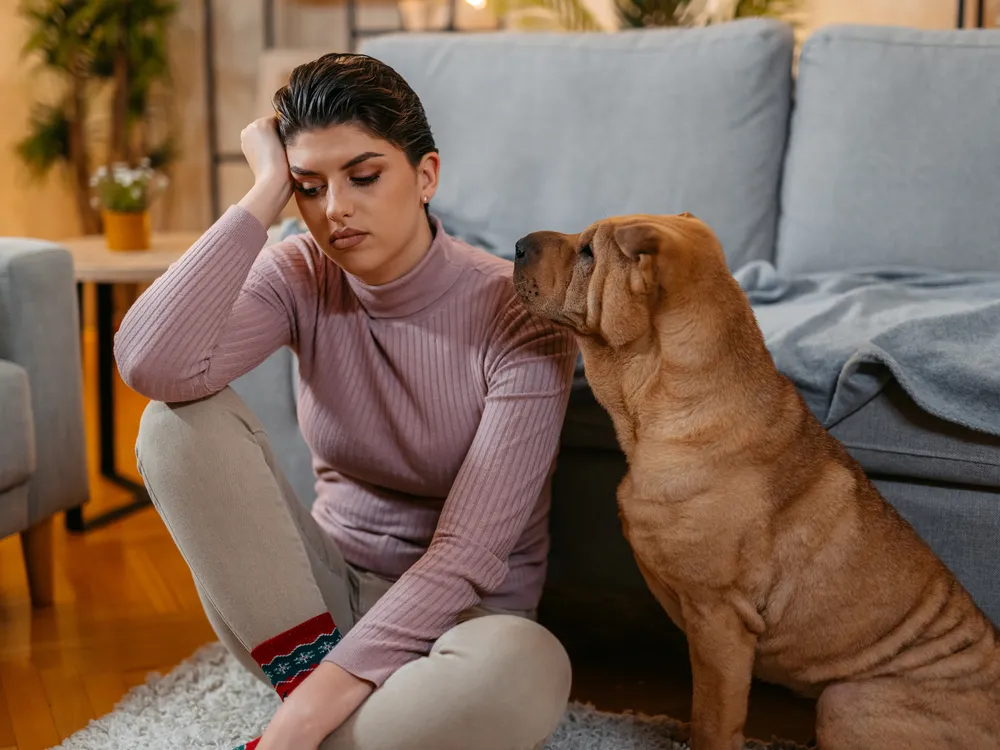 a woman sits on the floor in front of her couch, resting her elbow on her knee and looking sad, as her shar pei dog sits next to her, its head near her shoulder, and looks on attentively