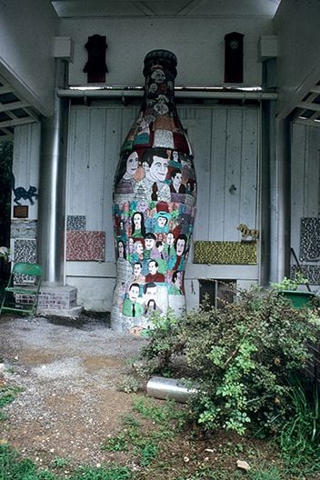 Large sculpture of a cola bottle painted with figures, standing next to a wall with other works of art displayed in a covered outdoor area with a bush in the right foreground.