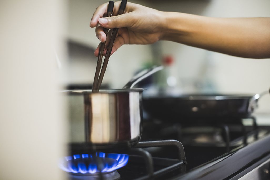 A woman's hand stirring a pot with chopsticks over a lit gas stove