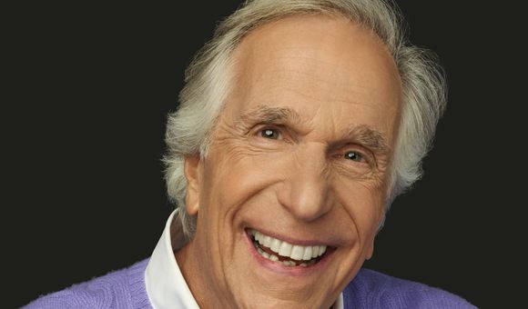 A headshot of Henry Winkler, shows a close-up the actor looking directly into the camera, his eyes twinkling above a broad, white smile.