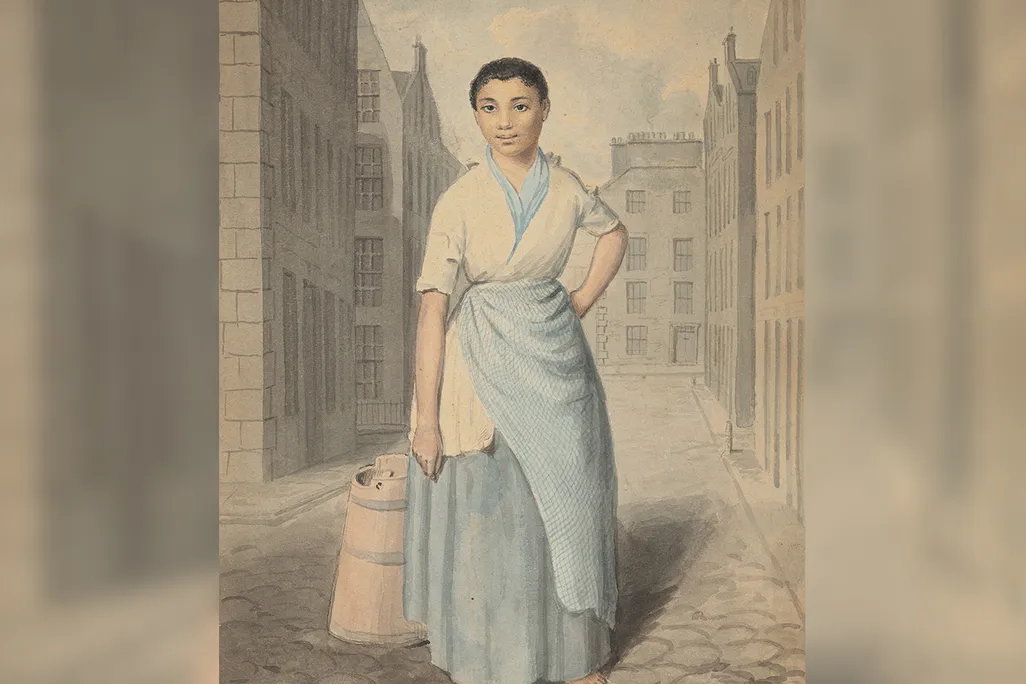 A solo full-length portrait of a Black woman in a white-and-blue wrapped dress, with cropped hair, standing with one hand on hip and barefoot on a cobbled street