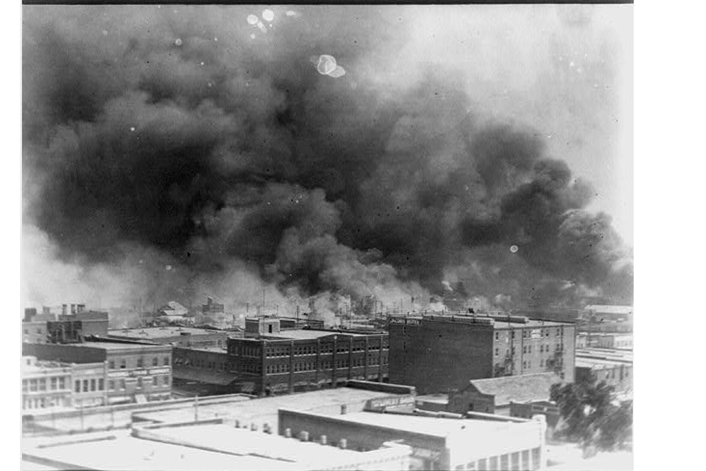 A black and white photo of buildings, from an aerial perspective, with thick plumes of black smoke rising above and obscuring the sky