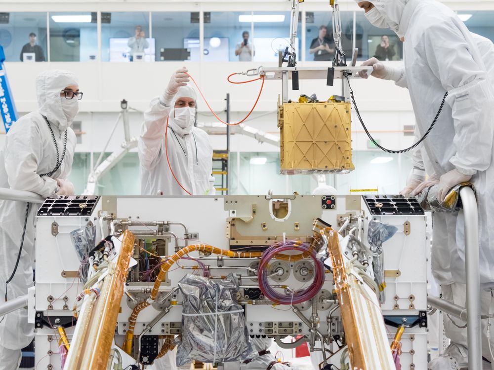 People in lab coats lower a gold box containing an oxygen-producing device into a Mars rover