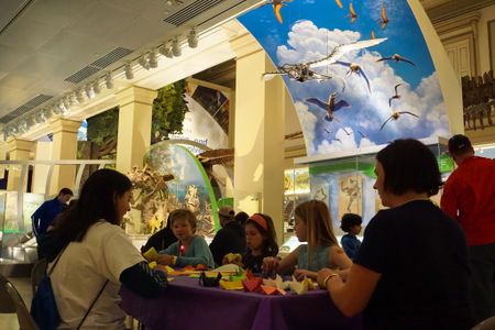 A group of children and two adults sit at a table creating origami birds among exhibits.