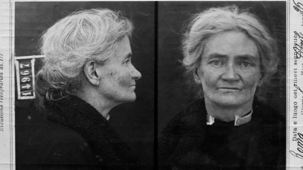 A black and white criminal mugshot of a white woman with graying hair, in a black outfit