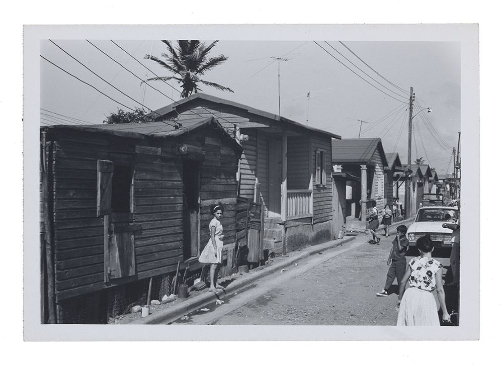 Black and white photograph of a street scene in Puerto Rico, with a girl standing on a curb wearing a light-colored dress with a full skirt.