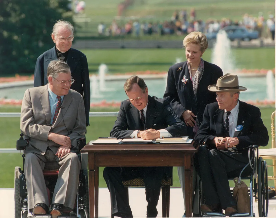 On a sunny day on the White House south lawn, President George Bush sits at a table and signs the 1990 Americans with Disabilities Act into law. On his right sits Evan Kemp, who uses a wheelchair. To his left, Justin Dart Jr., who wears a cowboy hat.