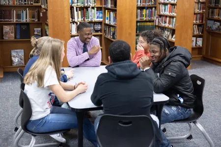 Teacher Kurt Russell sits at a square table in the school's library with five students, smiling and in conversation.