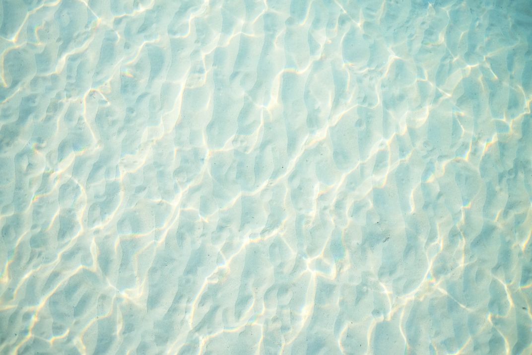 Translucent teal waters cast rippling shadows on an empty expanse of underwater sand.