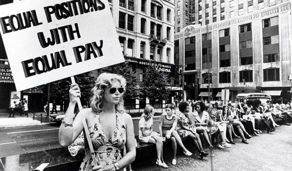 Woman holding a sign reading “Equal positions with equal pay” during an equal pay demonstration in a city plaza, with women seated behind her.
